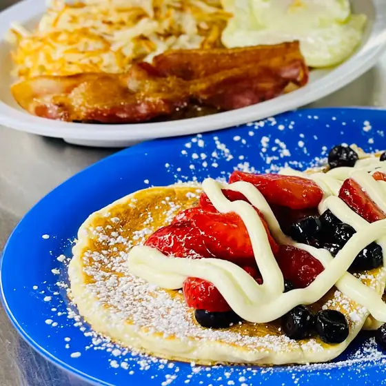 Pancake topped with strawberries, blueberries, a cream drizzle and powdered sugar on a blue plate, with bacon, eggs and hash browns behind